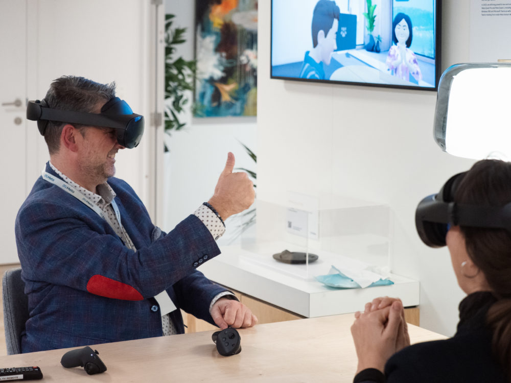 Mark Wheatley, a deaf middle aged person, uses a Virtual Reality set to sign to another person. In the background, we can see a screen with their avatars in the metaverse