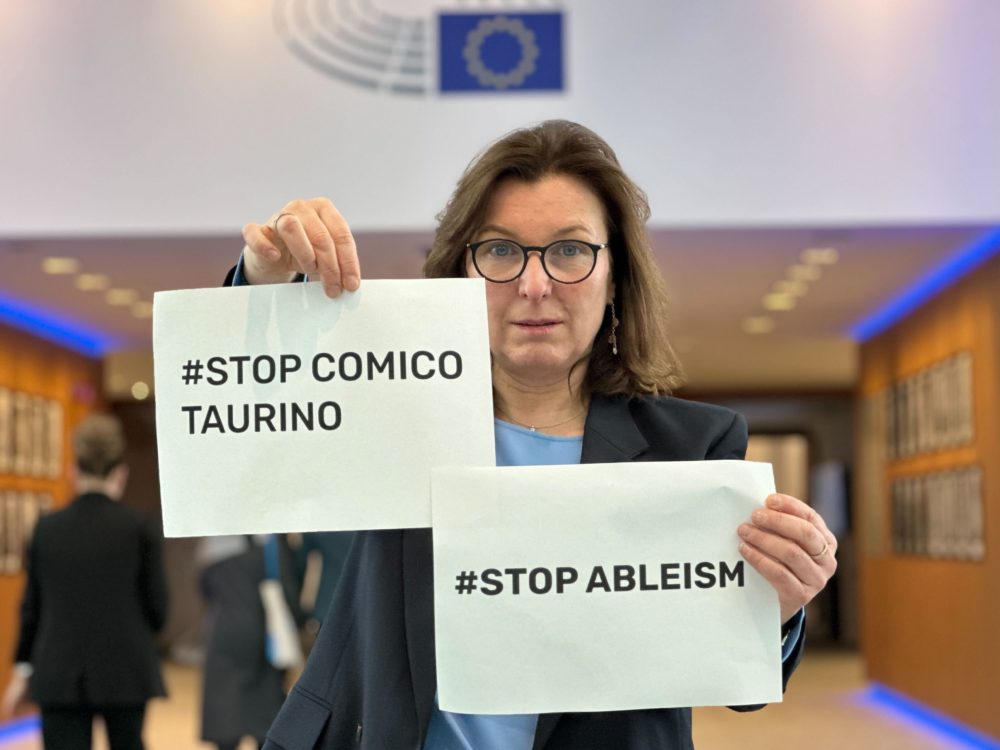 MEP Tilly Metz in the European Parliament, showing two signs. One says #StopComicoTaurino and one #StopAbleism