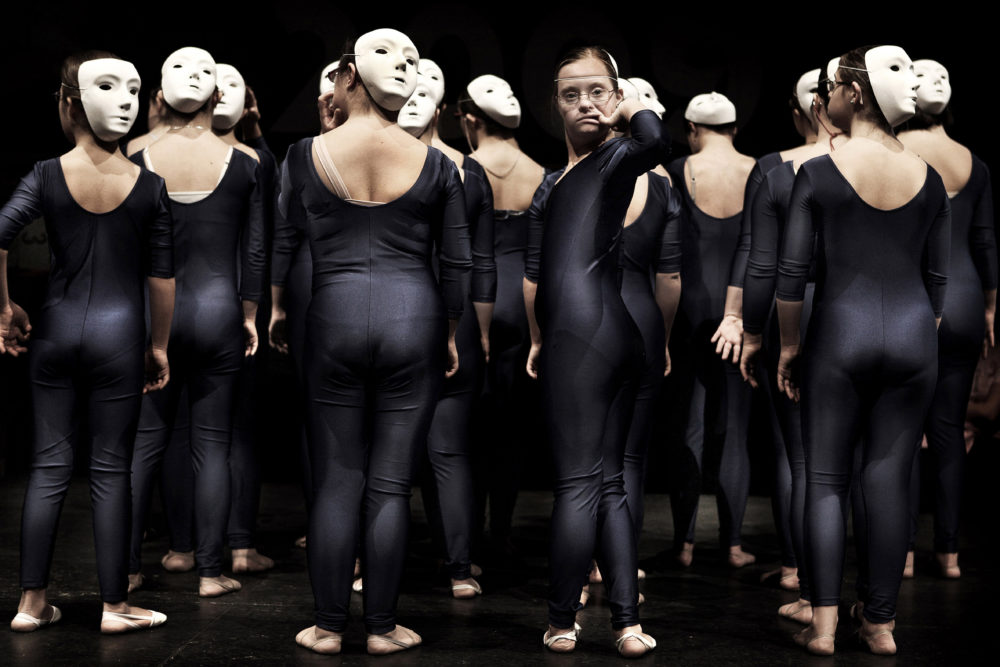 Artistic image with girls dressed in black tights with white masks on stage. All of them are on their backs, except for one who is a young woman with an intellectual disability.