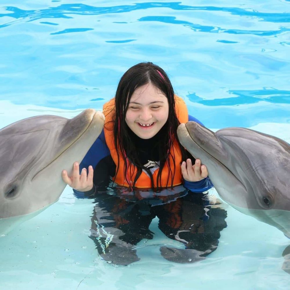 Image of a girl with Down syndrome in a swimming pool with a life preserver. Next to her are two dolphins. She smiles and touches them with her hands.