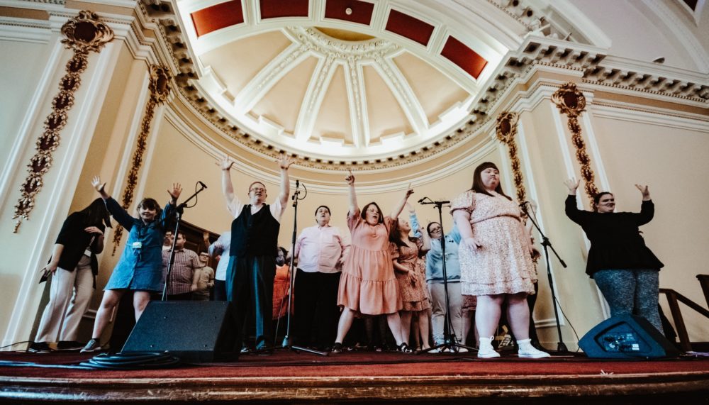 Image of a group of young musicians and singers with different disabilities. Some of them have their arms raised in joy. 