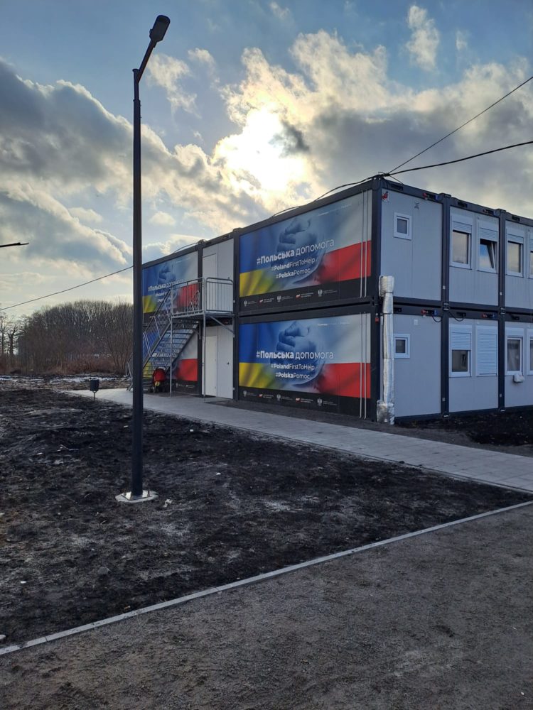 View of container homes, with an accessible footpath. The houses have an illustration of a mix of Ukranian and Polish flags saying "Poland first to help'