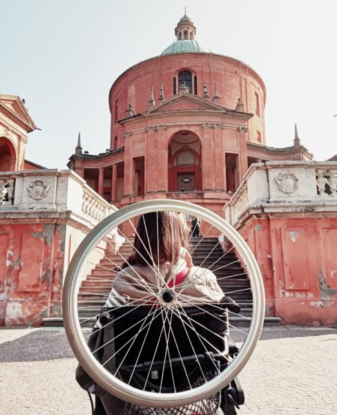 Wheelchair user, with a wheelchair wheel attached to the back of the wheelchair, faces steps to get to a churcj