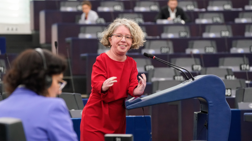 MEP Katrin Langensiepen, a short woman with a visible disability speaks from the podium. She wears a bright red dress.