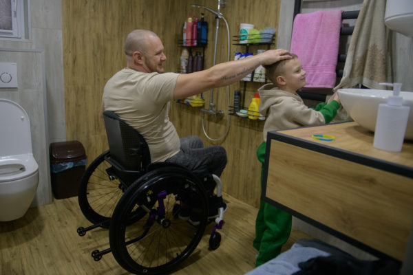 A middle aged man in a wheelchair stands in a spacious wooden bathroom pats the head of a young kid, likely is son, while the kid dries is hand in a towel. We notice a tilet and a sink that seem adapted to their heights.