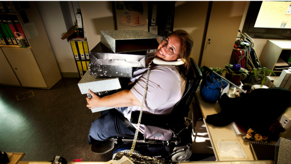 A middle aged women in a wheelchair smiles while she picks holds several folders and answers a phone in her office