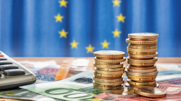 Piles of coins and banknotes are set on a table with an EU falg behind