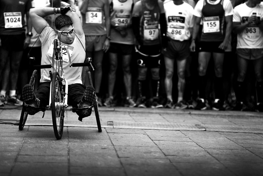 Adaptive sport: A young man is on an adapted bike, ready to start the race. Behind him is the starting line with the rest of the runners lined up
