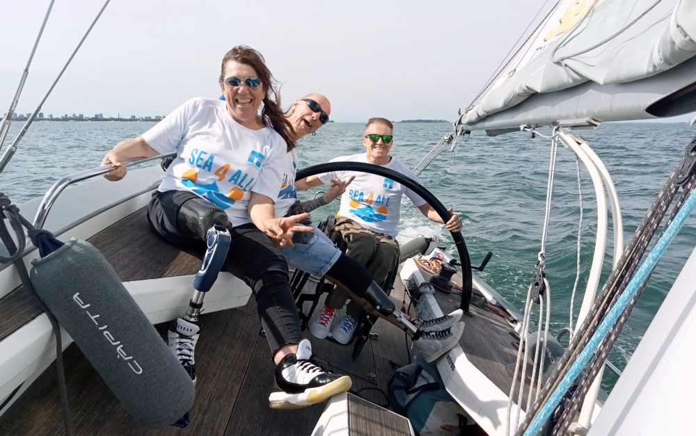 See4All - Adaptive sport. A group of friends with different physical disabilities (wheelchair users and one with a prosthetic) on a small sailboat, enjoying the sea and the sport. All three are smiling at the camera.