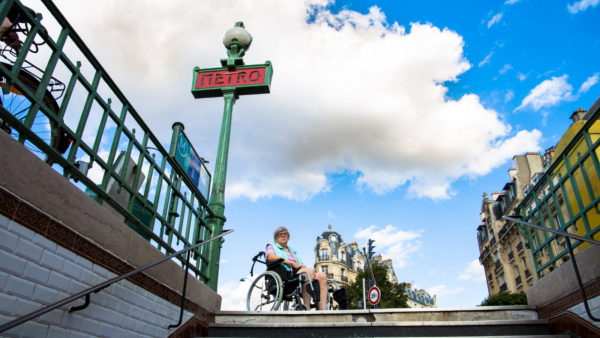 An older woman in a wheelchair facing a metro entrance that does not have a ramp or lift