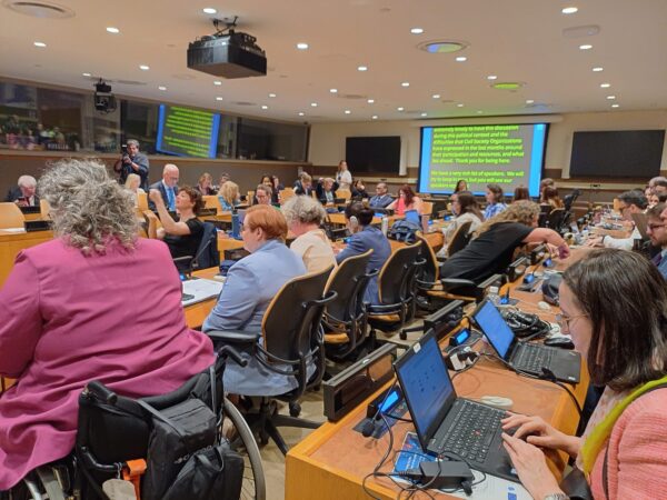 Conference room at the United Nations with a room filled with many attendees, facing a large screen displaying real time captionst. Participants engage, with one person raising a hand and another taking photos; interpretation facilities are visible.
